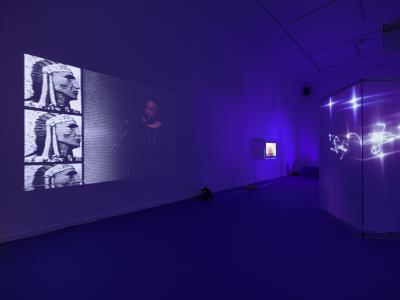An art gallery with a blue carpet containing several props, and lights. Blue light is cast upon the walls, with projected stars and abstract patterns adorning some of the props. In the foreground, a video of someone speaking into a microphone and a repeated photograph of an indigenous person in headdress is projected onto the wall.
