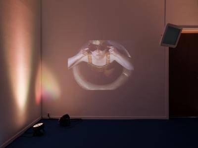 A video of a person in goggles and swimwear in a ring buoy is projected onto a wall cast in pink light. In the corner, two lights sit on a blue carpet. To the right of the projection is a small monitor suspended from the ceiling.