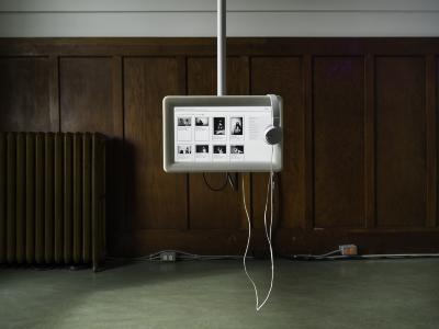 A pair of white headphones rest on a small monitor that is suspended from the ceiling. On the screen, there are several black and white images. In the background, a brass radiator sits against a wood-paneled wall.