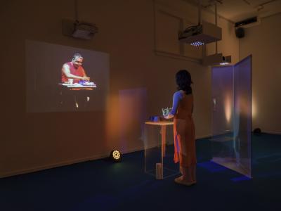 An exhibition guide wearing an orange apron stands holding a teapot over cups placed on a plexiglass plinth. On the wall opposite to her is a video projection of a man opening a lunchbox. The walls are lit in a soft red light, contrasting the deep blue of the carpet.