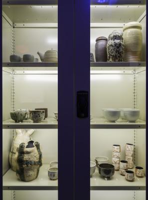 A close up shot of a shelving unit containing various ceramic vases, tea pots, teal bowls, and cups. Its doors are closed and it is illuminated from the inside with LED strips.
