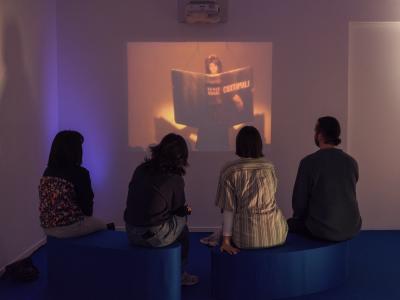 Four individuals sit on blue oval-shaped benches placed on a blue carpet. They are watching a video projection of a person holding and reading an oversized book titled CUXTAMALE.