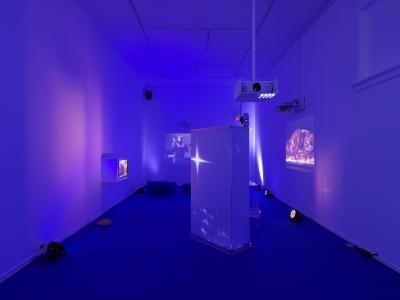 An art gallery with a blue carpet containing several props, lights, and projectors. Blue and magenta light is cast onto the walls. Also projected on the far side of the room, is a video of a man shaking a maraca, and on the right wall, a video of a bloodied body wrapped in plastic laying on top of a table.