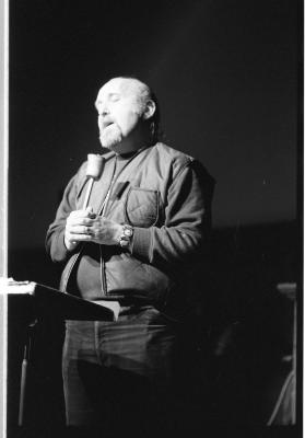 A man stands holding a microphone against his torso. His eyes are closed and head is tilted as he speaks into the mic. In front of him is a music stand. His face is cast in stark lighting that fades to the rest of his body. This image is in black and white.